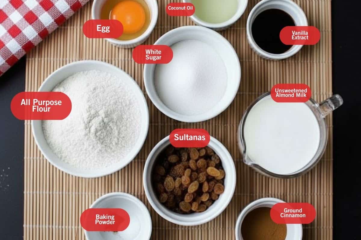 Flat lay photo of ingredient shot of all purpose flour, egg, coconut oil, white sugar, sultanas, baking powder, ground cinnamon, unsweetened almond milk and vanilla extract