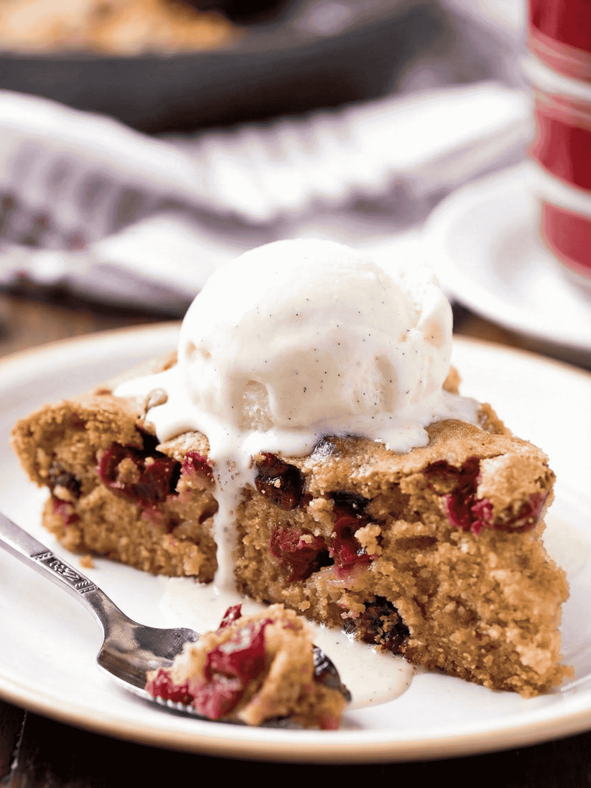 side shot of a slice of cobbler cake with cranberries and a scoop of vanilla ice cream on top melting over the slice and a spoon with a piece of cake on it sitting on a white plate - 1