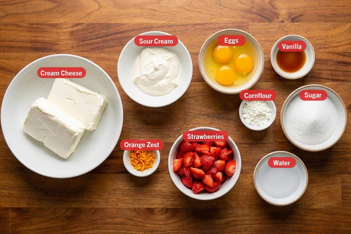 Top shot of ingredients: cream cheese, sour cream, eggs, vanilla, cornflour, sugar, orange zest, strawberries, and water.