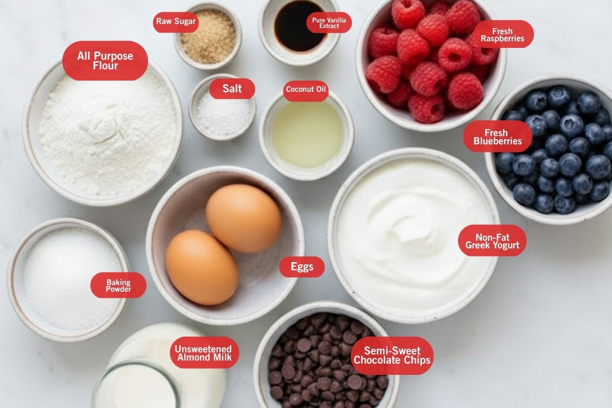 Flat lay photo of ingredient shot of all purpose flour, raw sugar, pure vanilla extract, fresh raspberries, fresh blueberries, semi-sweet chocolate chips, non fat greek yogurt, unsweetened almond milk, baking powder, coconut oil, salt