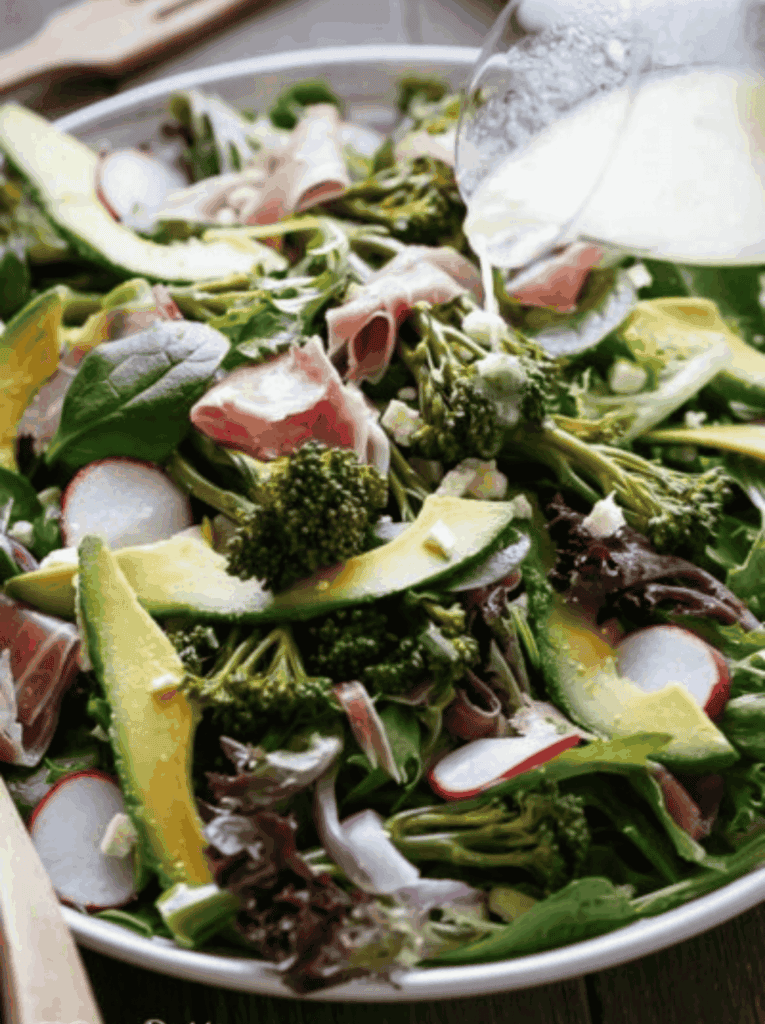 close up shot from top down showing a white bowl filled with broccolini salad with prosciutto, feta and avocado with a jug of dressing being poured onto the salad - 9