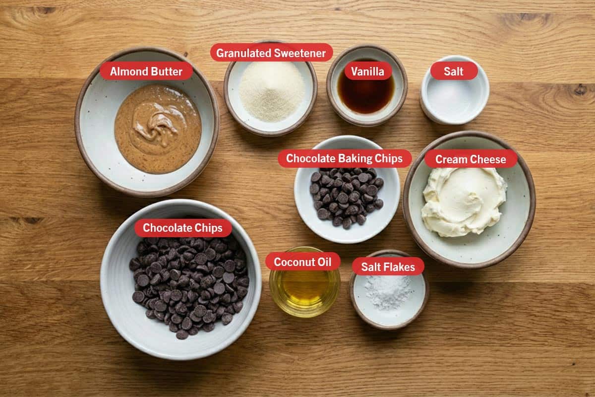 Top shot of ingredients: almond butter, granulated sweetener, vanilla, salt, chocolate chips, chocolate baking chips, cream cheese, coconut oil, and salt flakes.