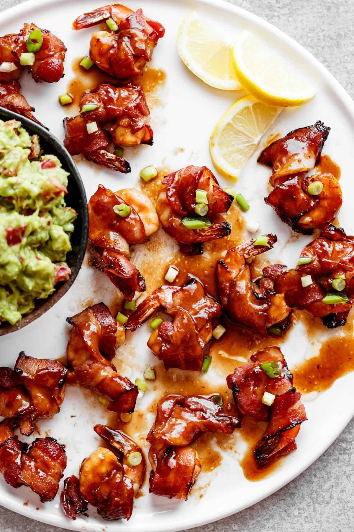 Crispy Bacon Wrapped Shrimp drizzled in a sticky Honey Garlic Sauce with a side of guacamole in a ramekin and three half slices of lemon. 