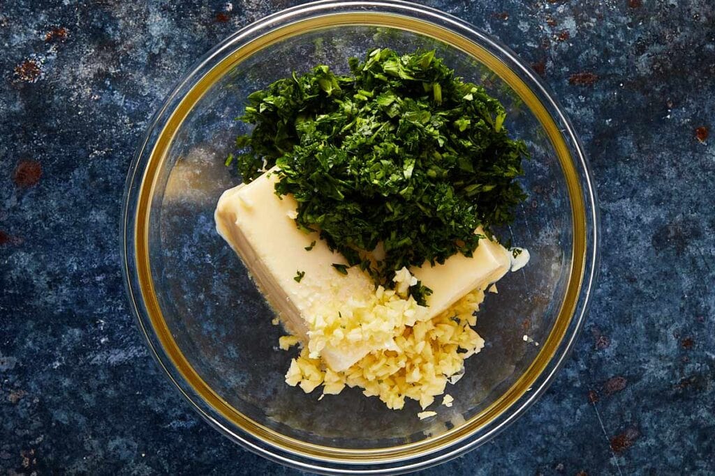 a top down shot of a glass bowl sitting on a blue dark stone bench with butter and fresh herbs inside