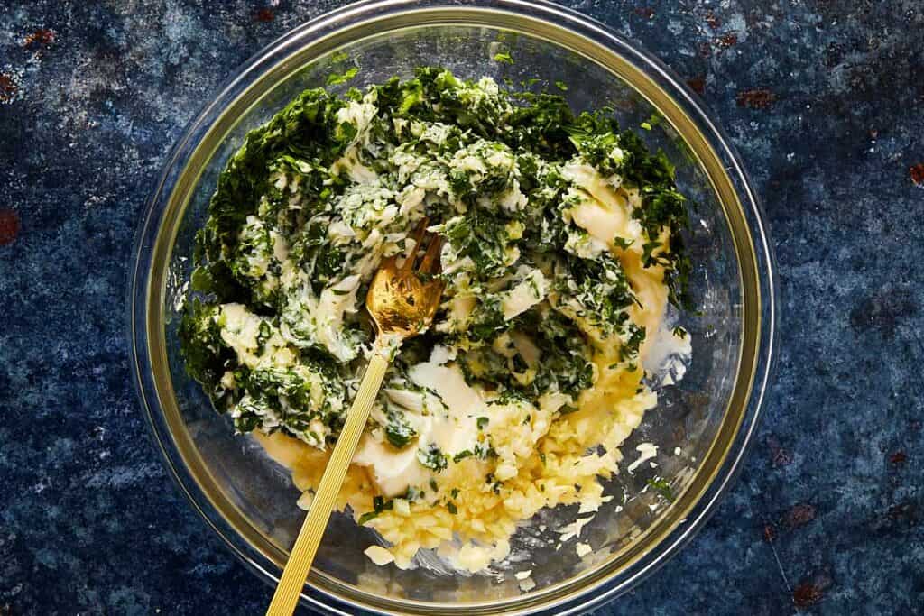 a top down shot of a glass bowl sitting on a blue dark stone bench with butter and fresh herbs inside with a gold spoon mixing the ingredients
