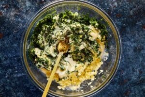 a top down shot of a glass bowl sitting on a blue dark stone bench with butter and fresh herbs inside with a gold spoon mixing the ingredients