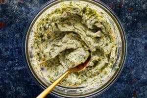 a top down shot of a glass bowl sitting on a blue dark stone bench with butter and fresh herbs inside with a gold spoon mixing the ingredients into a paste