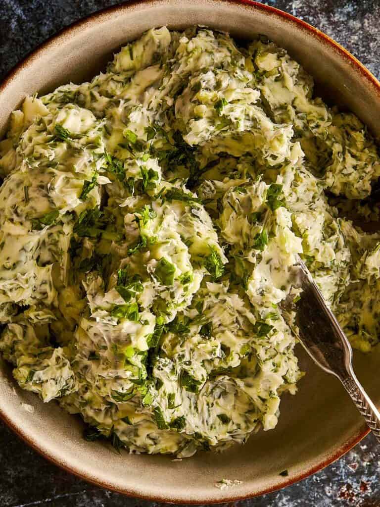 a top down shot very close up of a glass bowl sitting on a blue dark stone bench with butter and fresh herbs inside with a gold spoon mixing the ingredients into a paste
