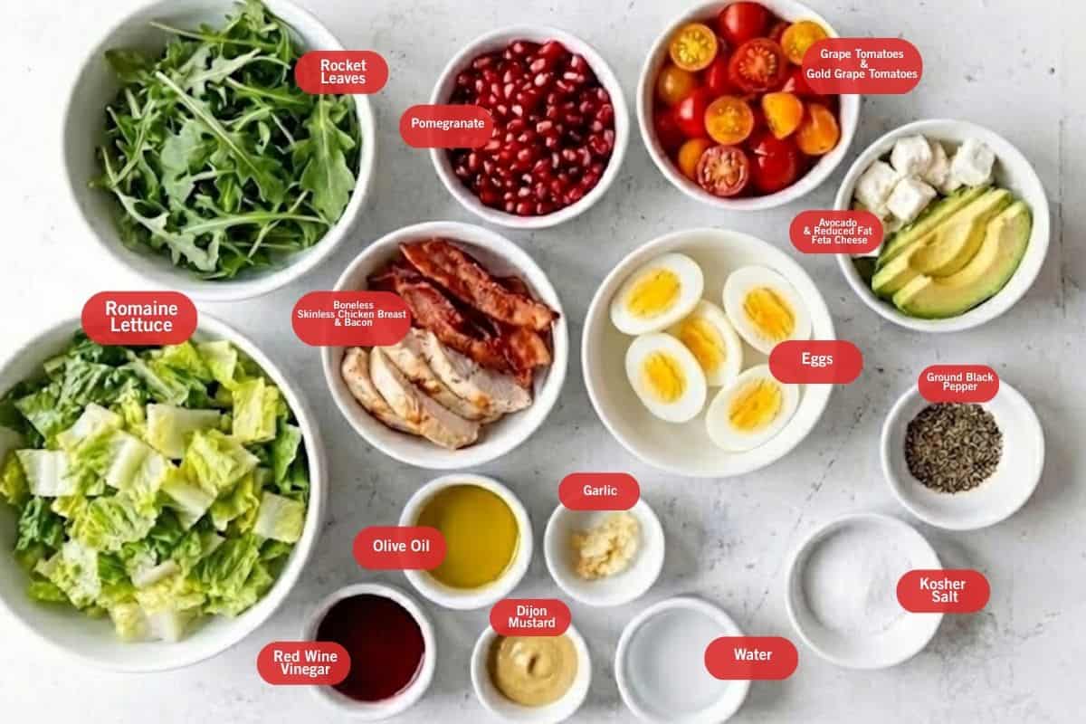 Flat lay photo of ingredient shot of Romaine lettuce, rocket leaves, pomegranate, grape tomatoes and gold grape tomatoes, avocado and reduced fat feta cheese, ground black pepper, kosher salt, water, garlic, dijon mustard, olive oil, red wine vinegar, boneless skinless chicken breast and bacon, eggs