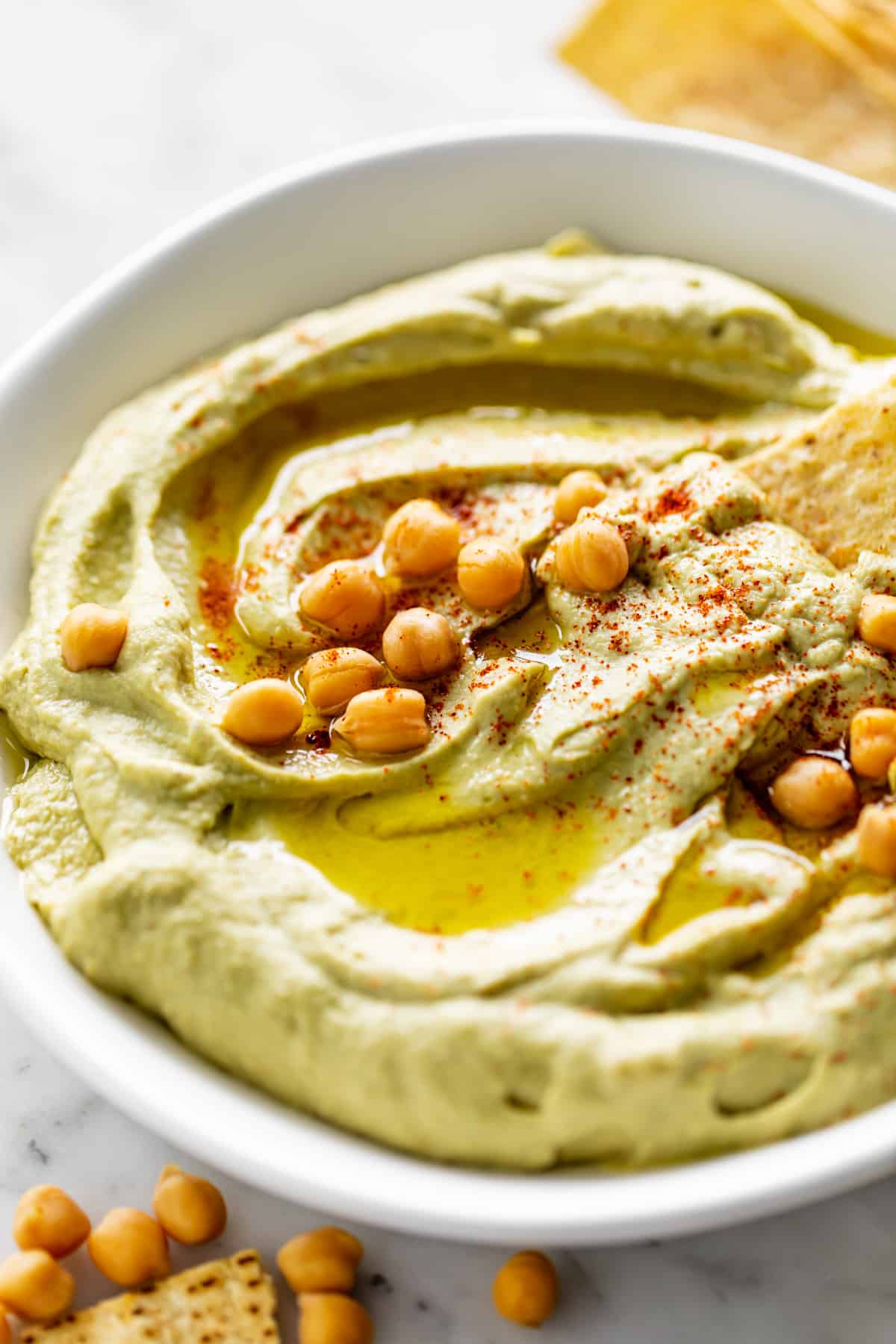 Avocado Hummus served in a white bowl, drizzled with olive oil and topped with 16 whole chickpeas. Whole chickpeas decorate the bottom left corner of the table with corn chips | cafedelites.com