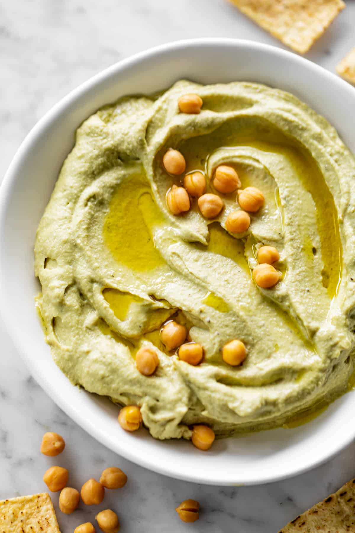 Avocado Hummus served in a white bowl, drizzled with olive oil and topped with 16 whole chickpeas. Whole chickpeas decorate the bottom left corner of the table with corn chips | cafedelites.com