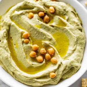 Avocado Hummus served in a white bowl, drizzled with olive oil and topped with 16 whole chickpeas. Whole chickpeas decorate the bottom left corner of the table with corn chips.