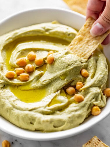 A corn chip dips into a white bowl full of Avocado Hummus, drizzled with olive oil and topped with whole chickpeas.