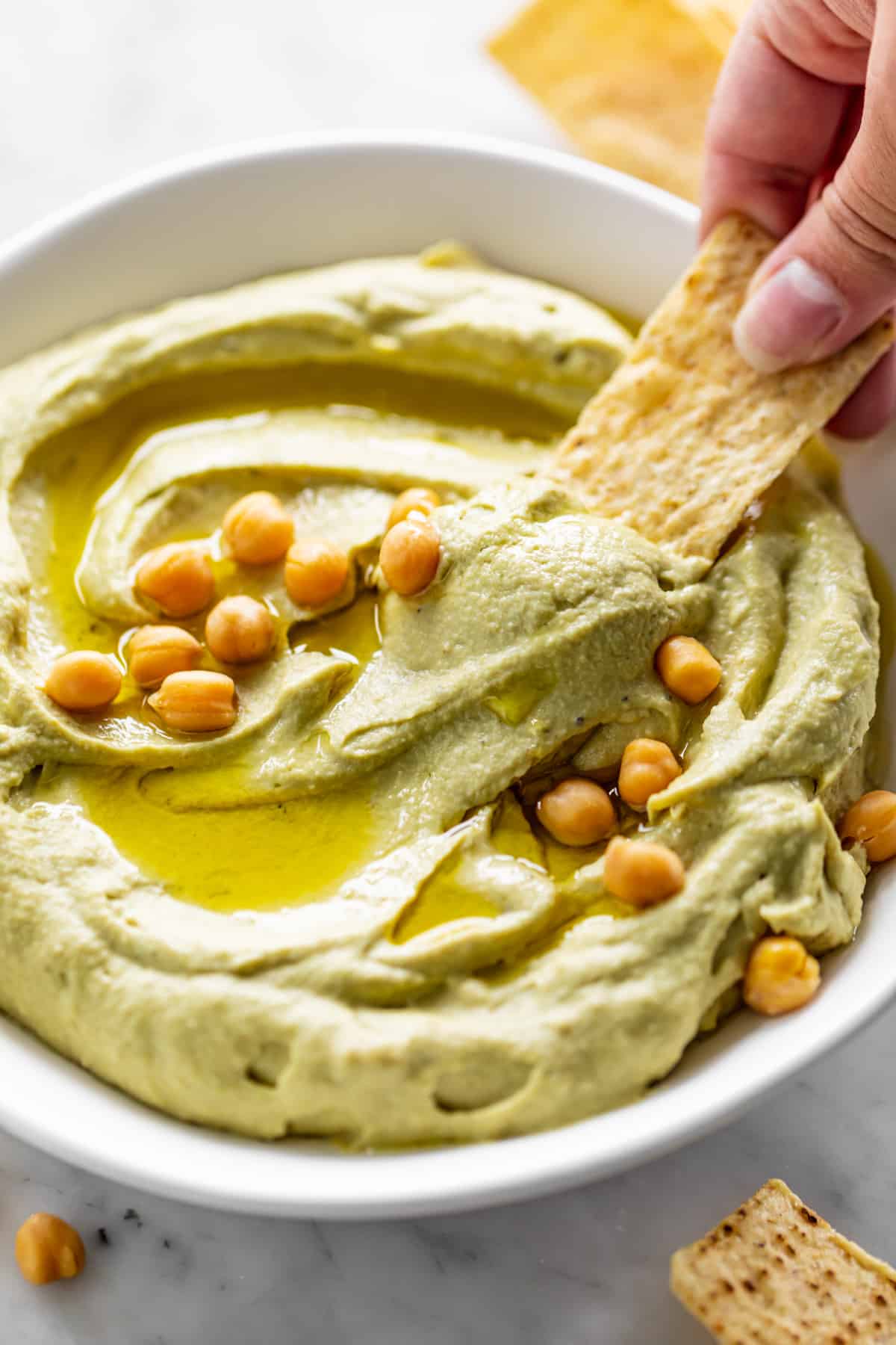 A corn chip dips into a white bowl full of Avocado Hummus, drizzled with olive oil and topped with whole chickpeas. | cafedelites.com