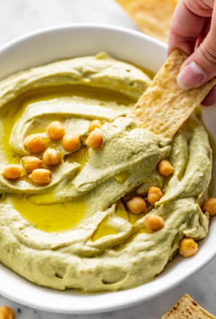 A corn chip dips into a white bowl full of Avocado Hummus, drizzled with olive oil and topped with whole chickpeas.