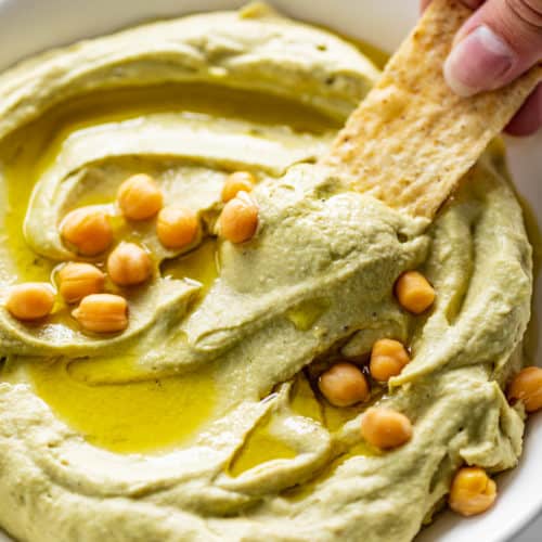 A corn chip dips into a white bowl full of Avocado Hummus, drizzled with olive oil and topped with whole chickpeas.