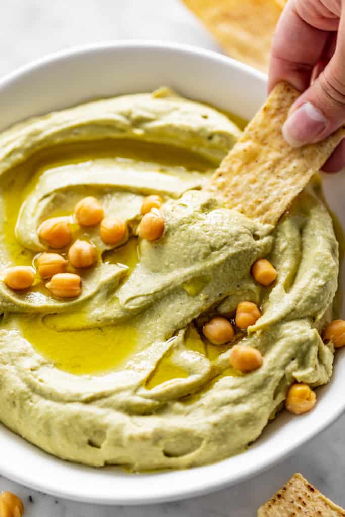 A corn chip dips into a white bowl full of Avocado Hummus, drizzled with olive oil and topped with whole chickpeas. | cafedelites.com