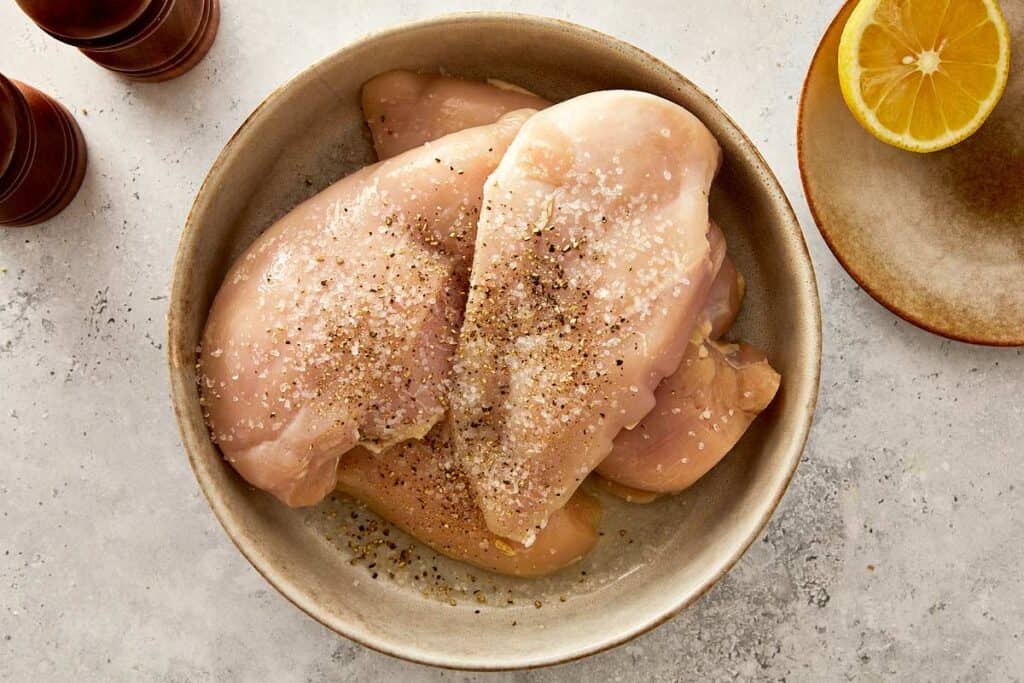 raw chicken breast salt and pepper on top in a white bowl with a lemon on the side
