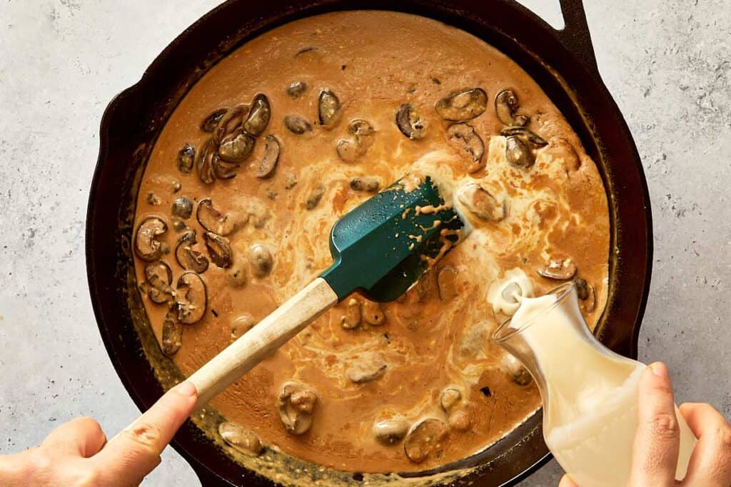 pouring some evaporated milk into a black skillet with brown sauce and mushrooms
