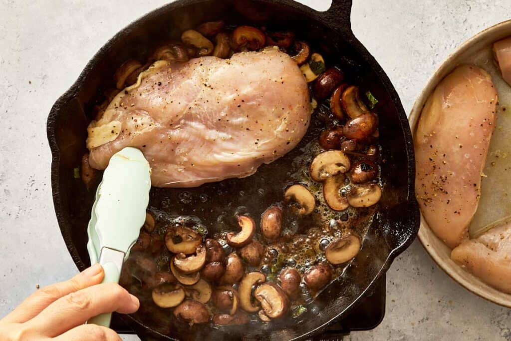 raw chicken breast being cooked in a skillet with mushrooms on one side and a pair of tongs lifting the chicken breast to see cooking