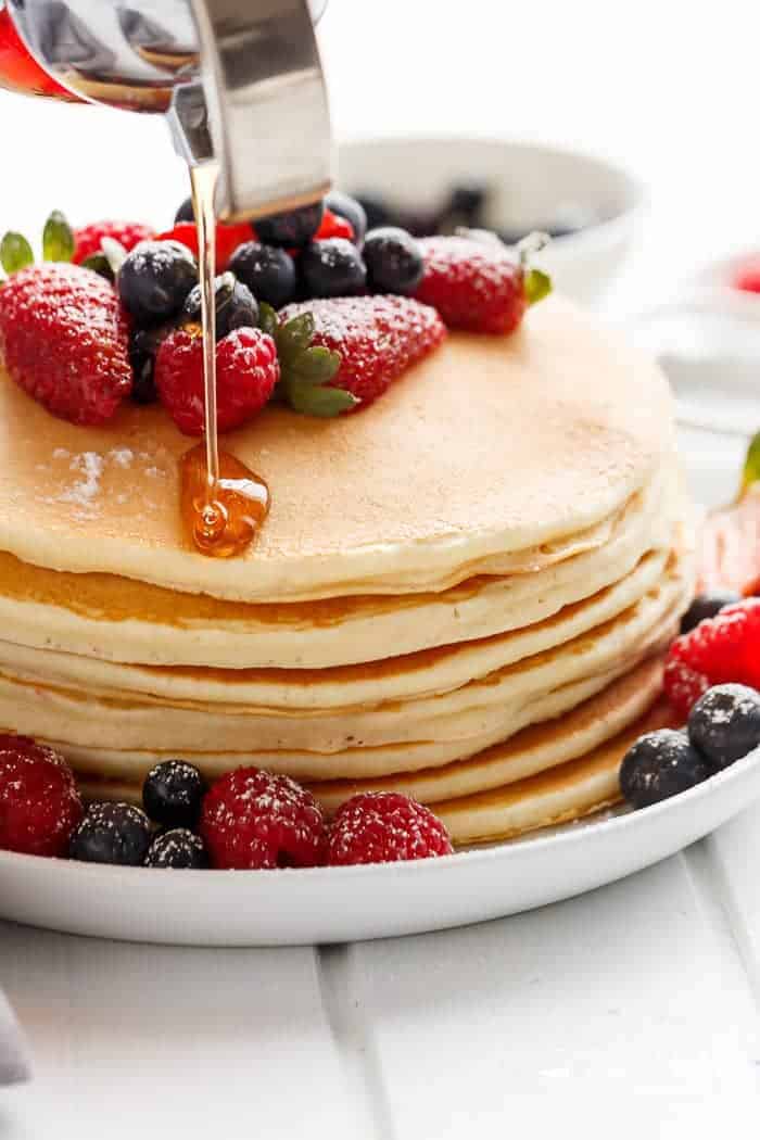 Maple syrup being drizzled over a large stack of pancakes with strawberries, blueberries and icing on top and around the sides. 