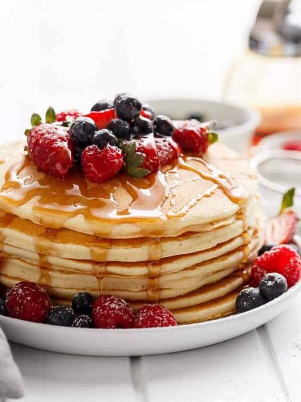 Close-up of a stack of pancakes drizzled with maple syrup and garnished with mixed-berries, cascading from the top to the bottom of the stack.