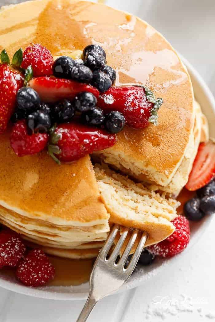 Bird's eye view of fork taking a large triangle chunk from a stack of pancakes. Blueberries and strawberries drizzled with maple syrup and icing sugar sit on top of the stack. 