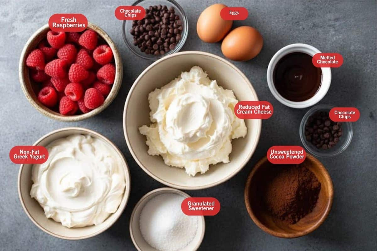 Flat lay photo of ingredient shot of fresh raspberries, chocolate chips, eggs, melted chocolate, chocolate chips, unsweetened cocoa powder, granulated sweetener, reduced fat cream cheese, on-fat greek yogurt
