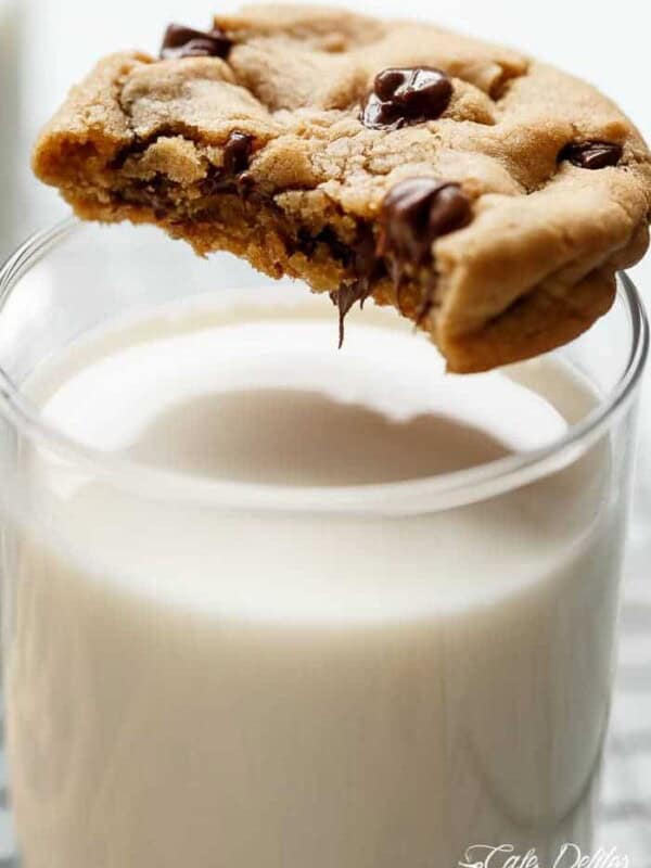 A close-up shot of a chocolate chip cookie being dipped into a glass of milk. The cookie is thick and chewy, with visible chocolate chips throughout. A bite has been taken out of the cookie, revealing a soft and gooey interior. The glass of milk is tall and clear, filled almost to the top with creamy white milk. The cookie is positioned over the glass, with the bitten portion submerged in the milk. In the background, two other chocolate chip cookies are partially visible, resting on a wire cooling rack.