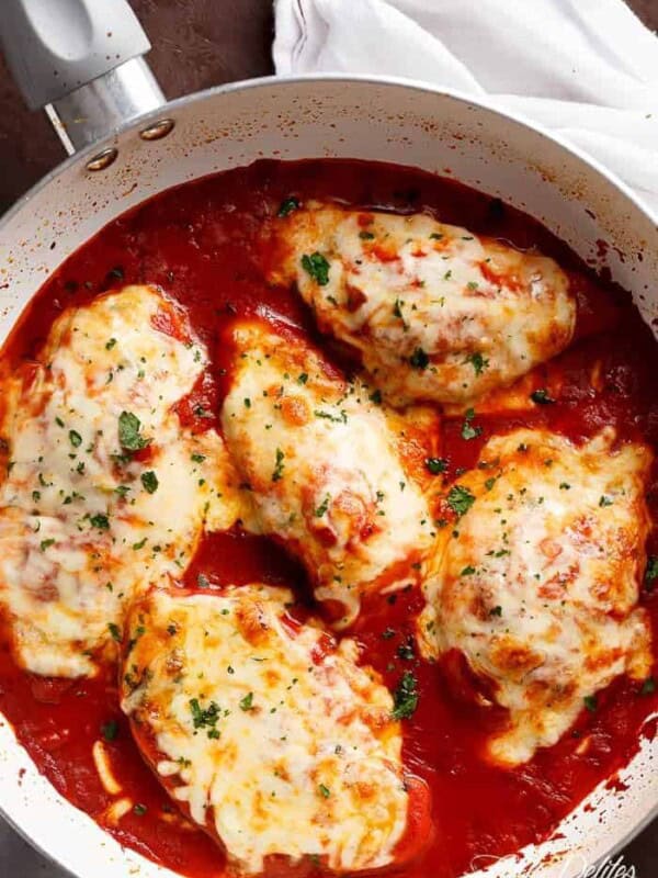 Five chicken breasts pieces, covered in grilled cheese, sprinkled with herbs, on a bed of tomato sauce in a white pan.