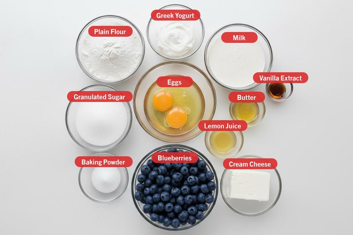 Top shot of ingredients: plain flour, greek yogurt, milk, vanilla extract, granulated sugar, eggs, butter, lemon juice, baking powder, blueberries, and cream cheese.
