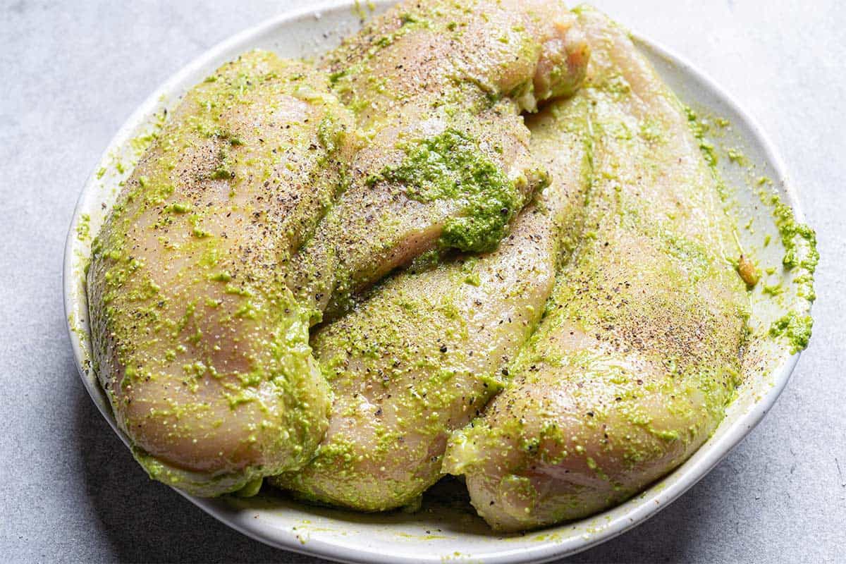 chicken breast in a white bowl being marinated in pesto