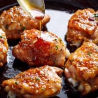 This close-up shot shows pan-seared chicken thighs coated in a vibrant orange glaze being drizzled with more of the glossy sauce from a spoon. The chicken, cooked to a delicious golden brown, glistens under the light, its surface sprinkled with herbs and spices. The dark background enhances the visual appeal of the food, emphasizing its rich color and texture.