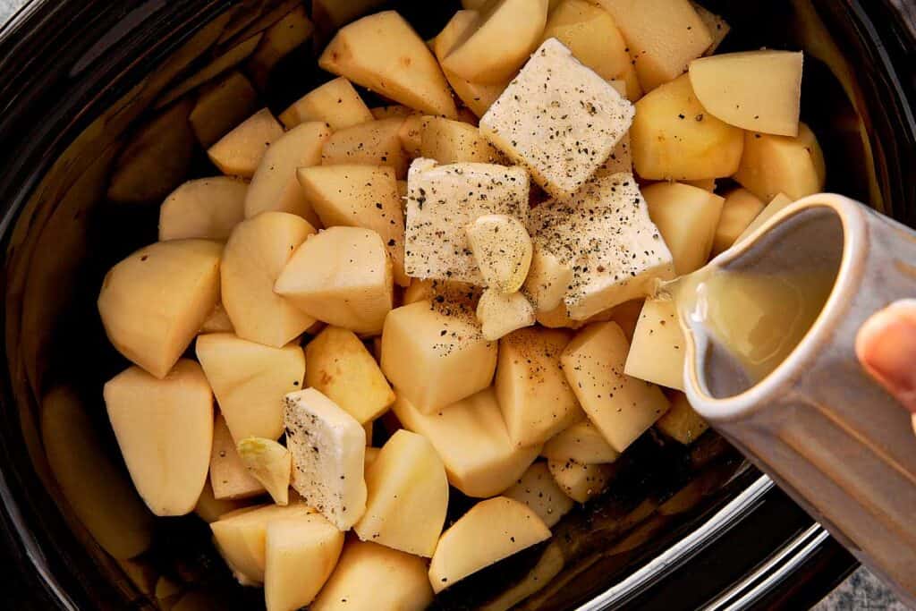 a top down view of cut up in cubes white potatoes in a slow cooker with butter cubs inside and a jug of milk being poured in - 5