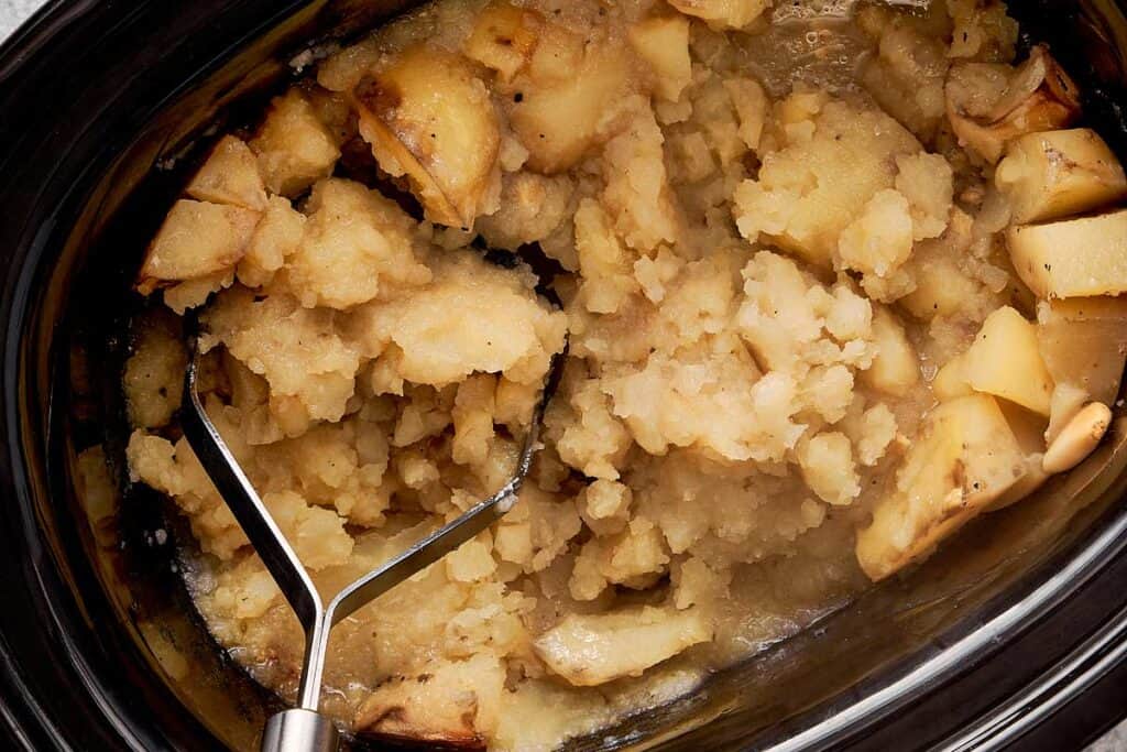 a top down view of baby potatoes in a slow cooker being mashed with a potato masher - 7