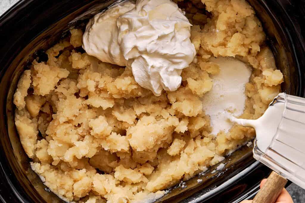 a top down shot of creamy potaotes already mashed and a dollop of cream on top and pouring milk in a slow cooker - 8