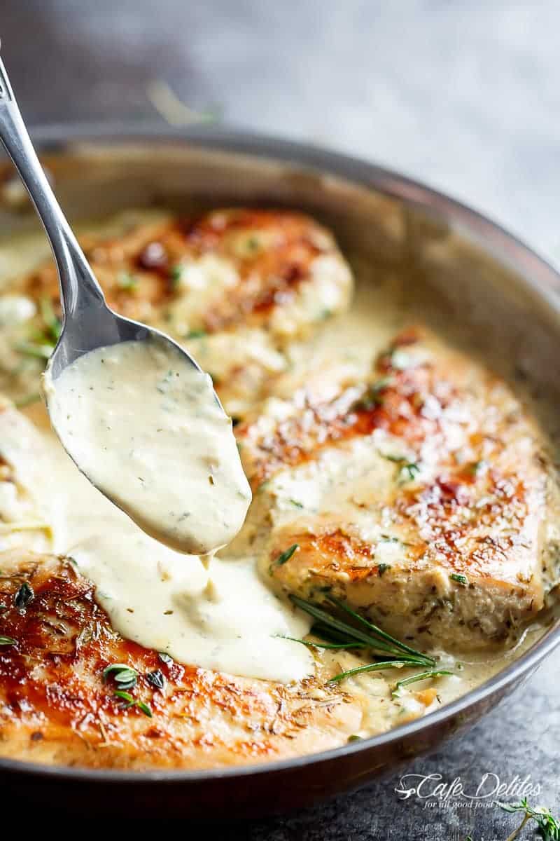 Thick sauce being poured over chicken in a pan, with a tablespoon. Fresh herbs including sprigs of rosemary sit on top of the chicken. 