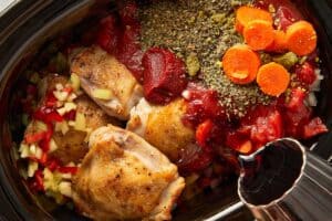 Chicken thighs layered over potatoes with vegetables, tomatoes, and seasonings in a slow cooker while Red wine being poured into slow cooker chicken cacciatore ingredients