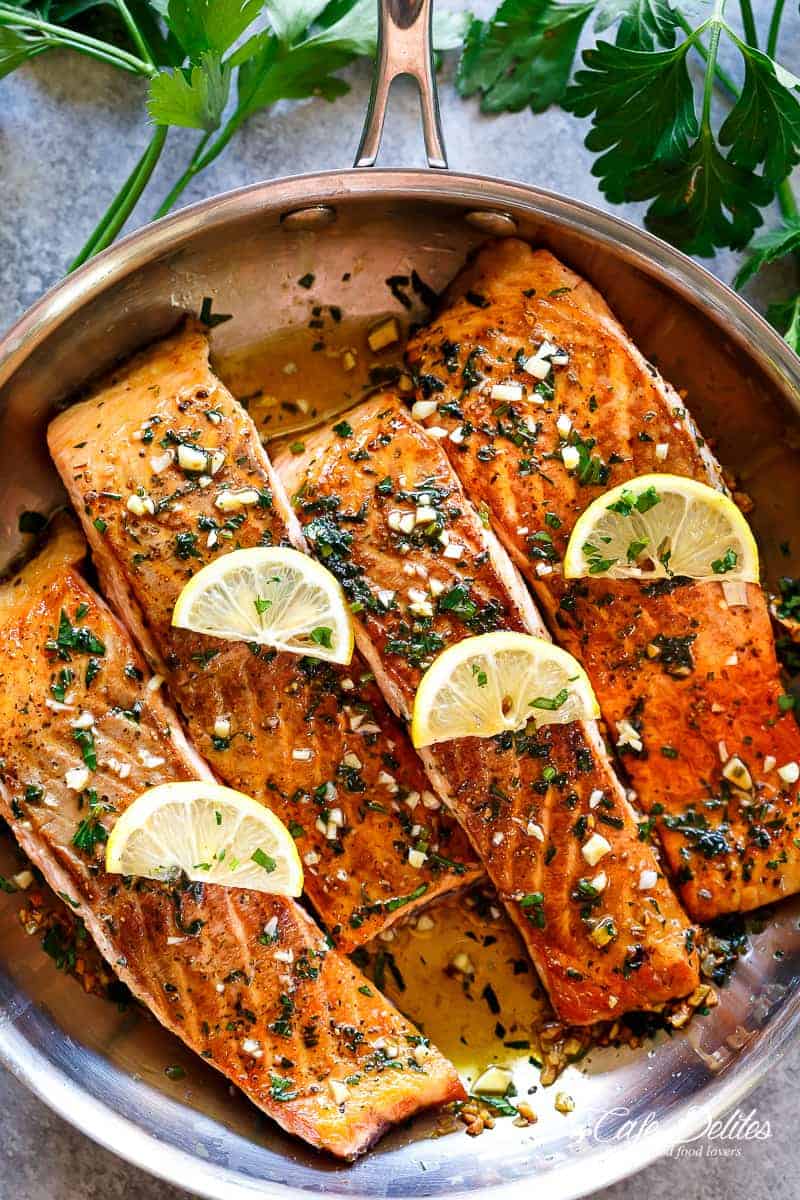 Four pieces of salmon in a pan with cooked herbs, garlic, melting butter and lemon slices on top. 