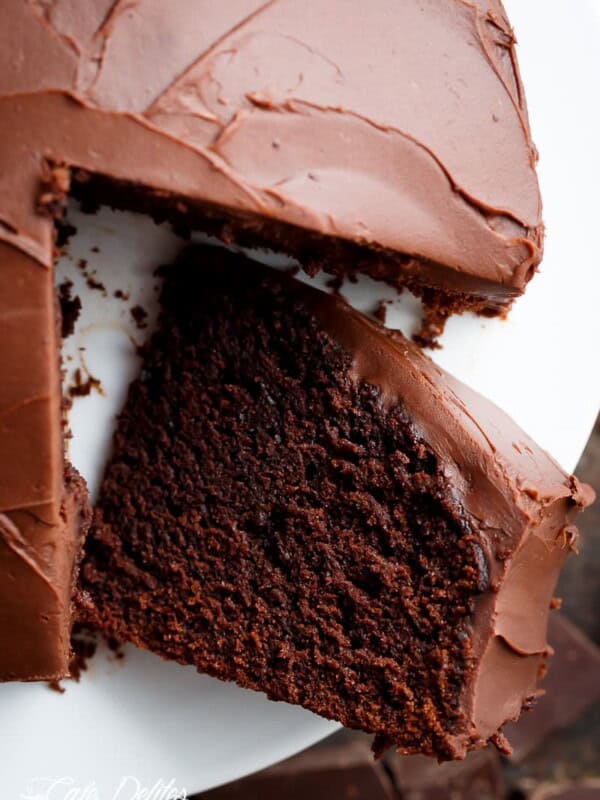 A rich fudgy chocolate cake, frosted in a decadent chocolate frosting, is shown sitting on a white plate. The first slice of cake has been cut lifted up from the rest of the cake. The chocolate frosting is smooth and shiny, while the cake is dense and moist looking. A pieces of chopped, chocolate cake is laying white plate to display the moist fluffy inside.