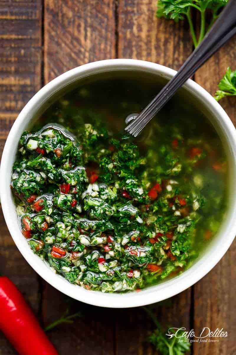 Top view of a bowl of Chimichurri with a sliver spoon, placed next to a chilli on a wooden bench.