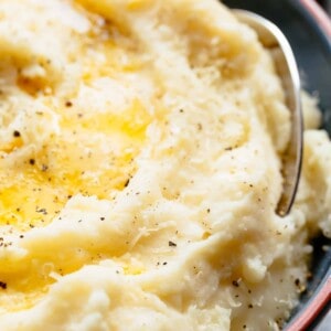 Close up image of Fluffly, mashed cauliflower in a shallow red and blue bowl with melted butter and salt and pepper in the centre being scooped with a silver spoon.