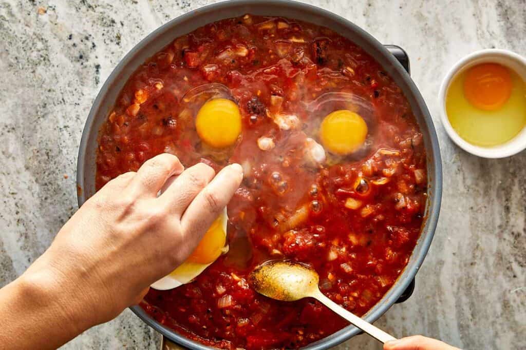 a top down view of a pan filled with diced vegetables and thick tomato salsa with two egg yokes raw cooking in the top and a spoon mixing with one hand and the other hand is adding in a bowl of sauce - 9