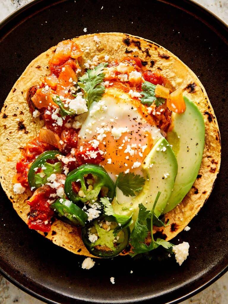 a top down view of a black plate with a large corn tortilla with eggs and tomato salsa and avocado in the middle - 11