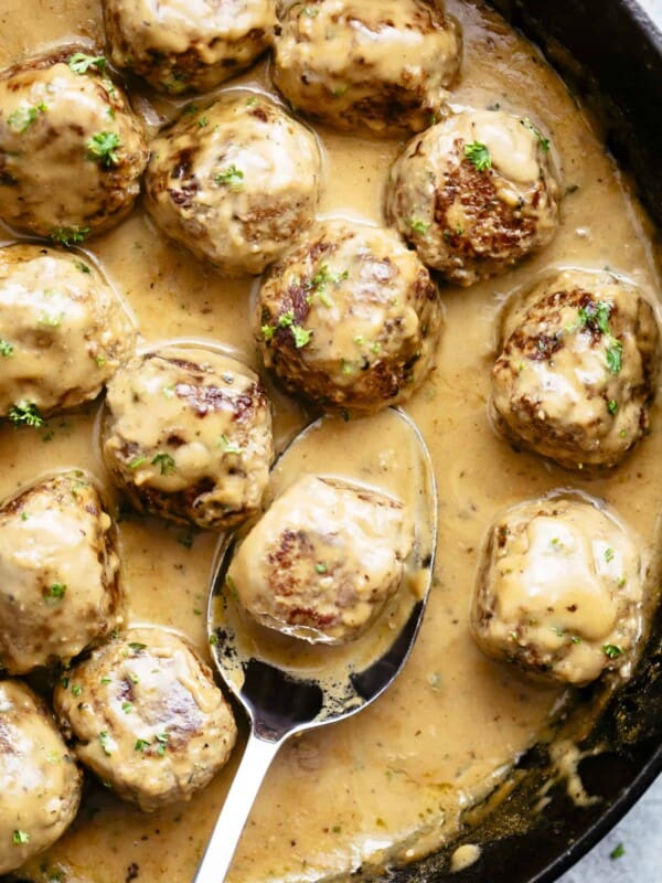 A top view of a cast iron skillet brimming with Swedish meatballs bathed in a creamy, beige gravy. The meatballs are browned to a rich, golden hue and generously speckled with fresh herbs. The skillet rests on a textured dark brown surface, hinting at the warmth of the dish.