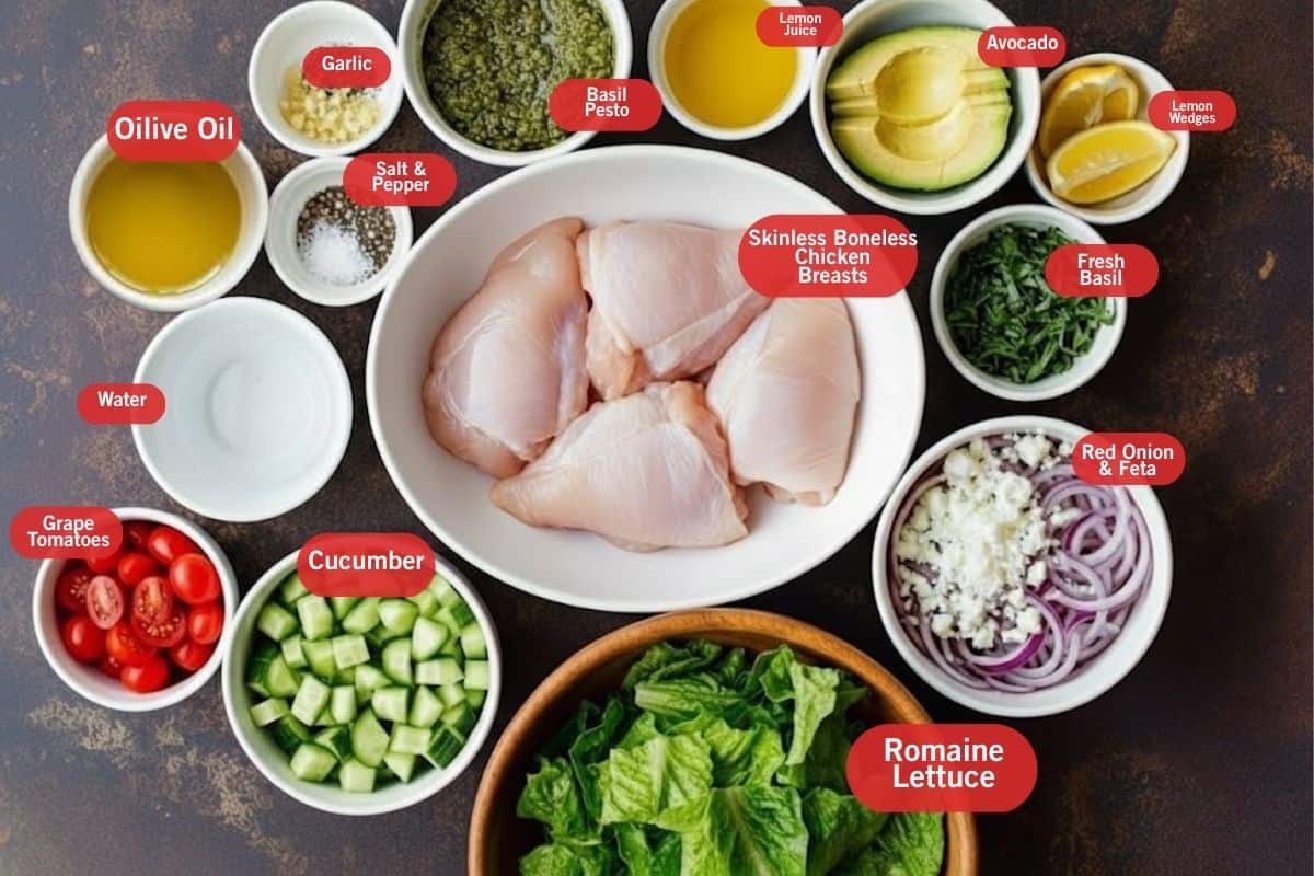 Flat lay photo of ingredient shot of olive oil, garlic, basil pesto, lemon juice, avocado, lemon wedges, fresh basil, red onion and feta, romaine lettuce, skinless, skinless boneless chicken breasts, cucumber, water, salt and pepper