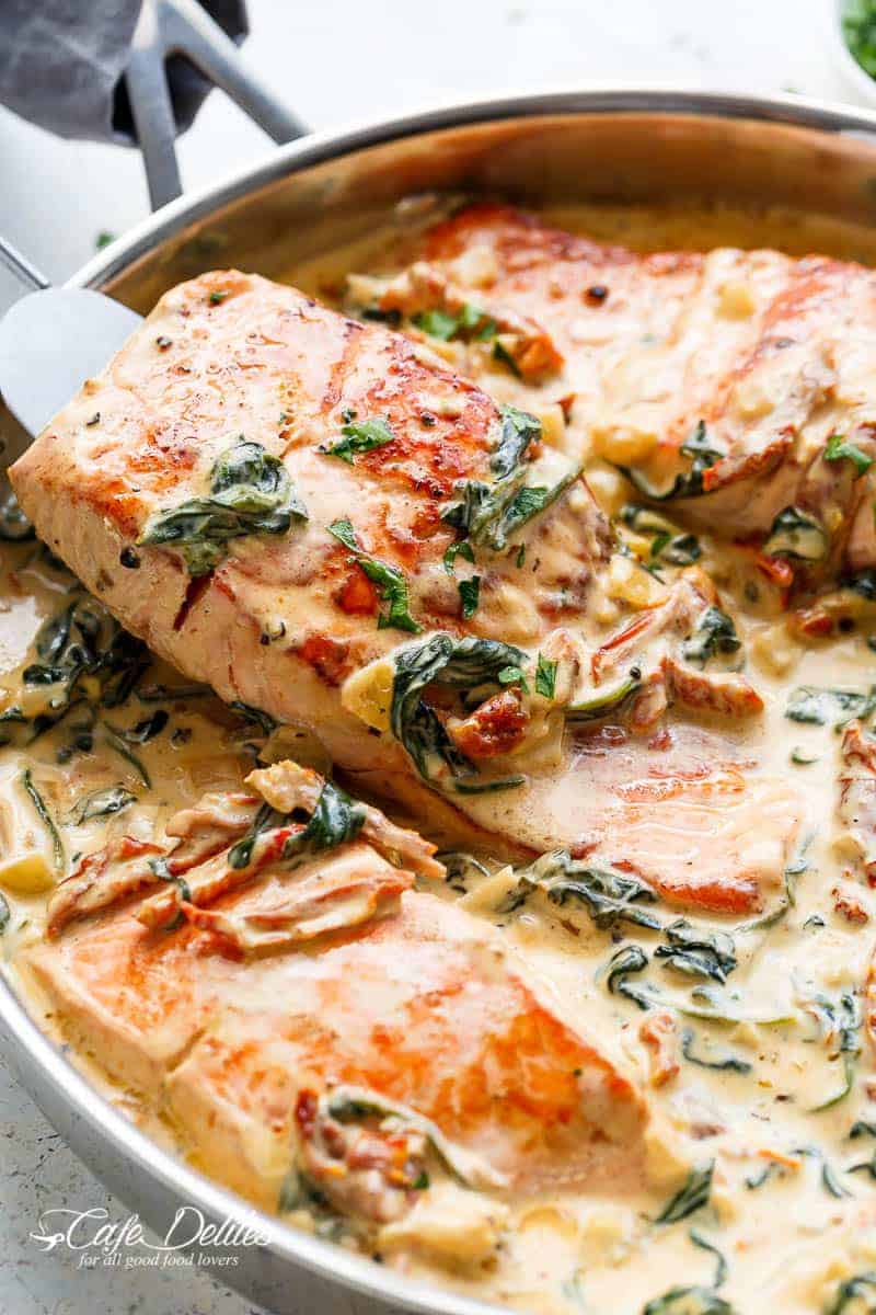 A silver spatula picks up a fillet of salmon simmered in a Creamy Tuscan style sauce with wilted spinach, sun dried tomato strips and basil strips. Salmon is presented in a silver frying pan against a white background | https://cafedelites.com