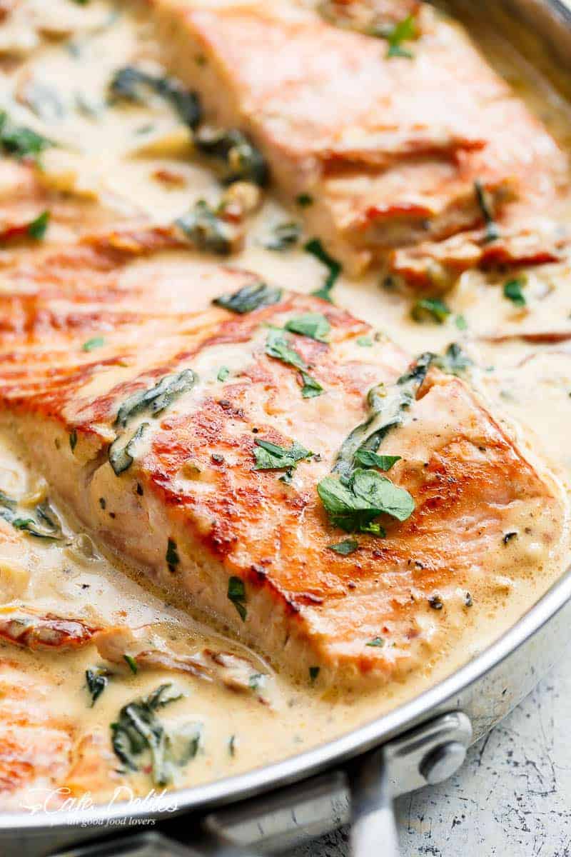 A close up of a piece of salmon in creamy Tuscan style sauce garnished with parsley. Salmon is presented in a silver frying pan with a white paint-scratched background. | https://cafedelites.com