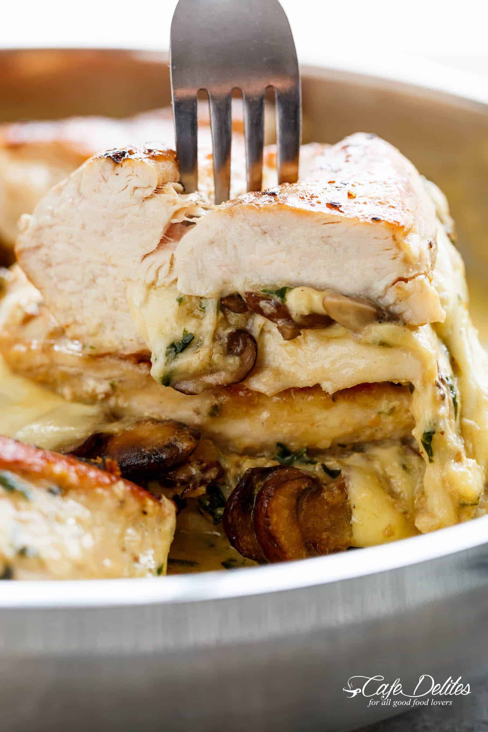 A fork pierces through cheesy mushroom stuffed chicken cut open! The piece is stacked on another stuffed chicken breast WITH the optional cream sauce in a silver frying pan.