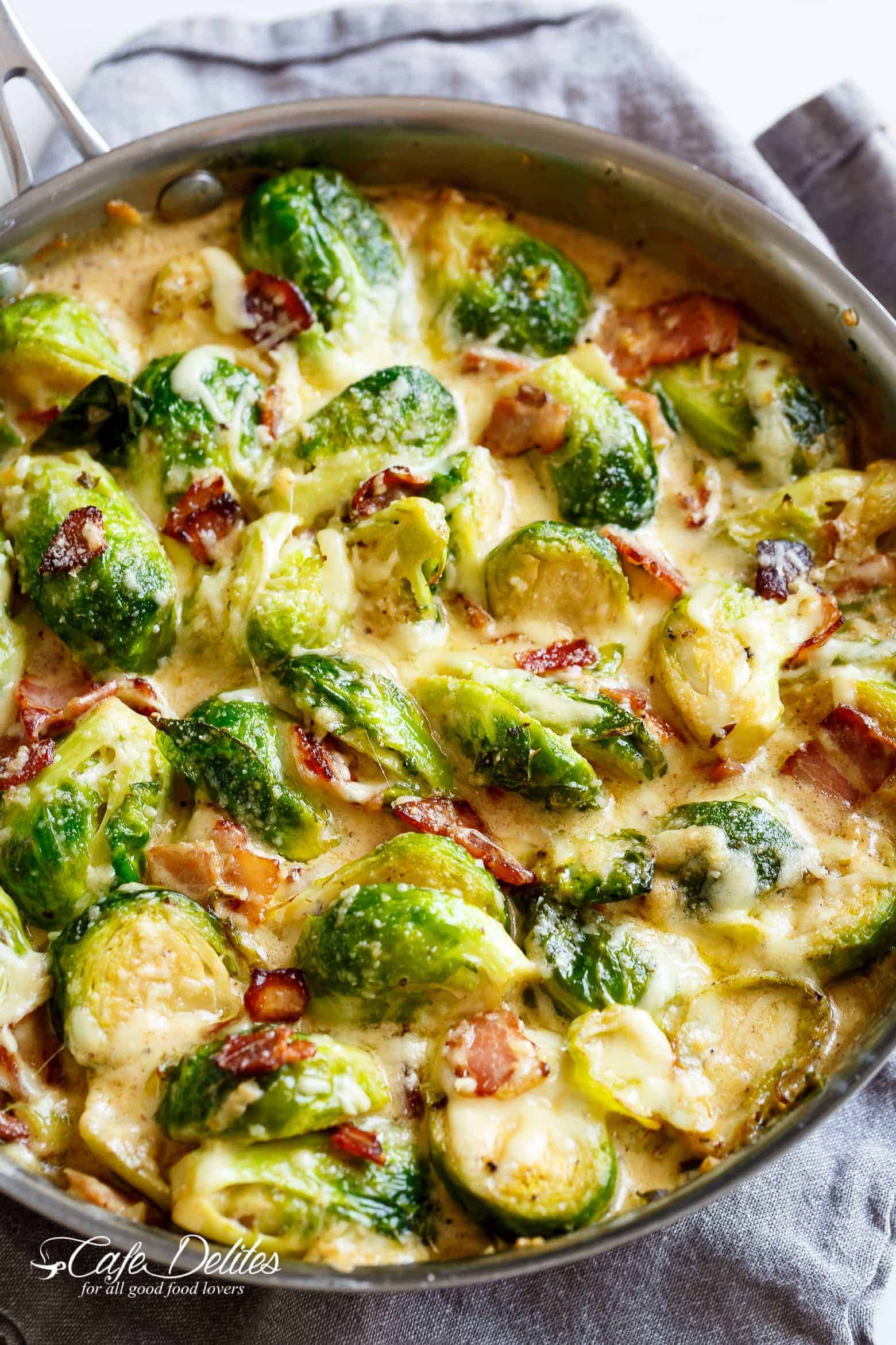 Brussel Sprouts with Bacon in a cheesy creamy garlic sauce, topped with bubbling mozzarella and parmesan cheese! Sprouts cooked in a silver frying pan, sitting on a grey tea towel on a white marble background. 
